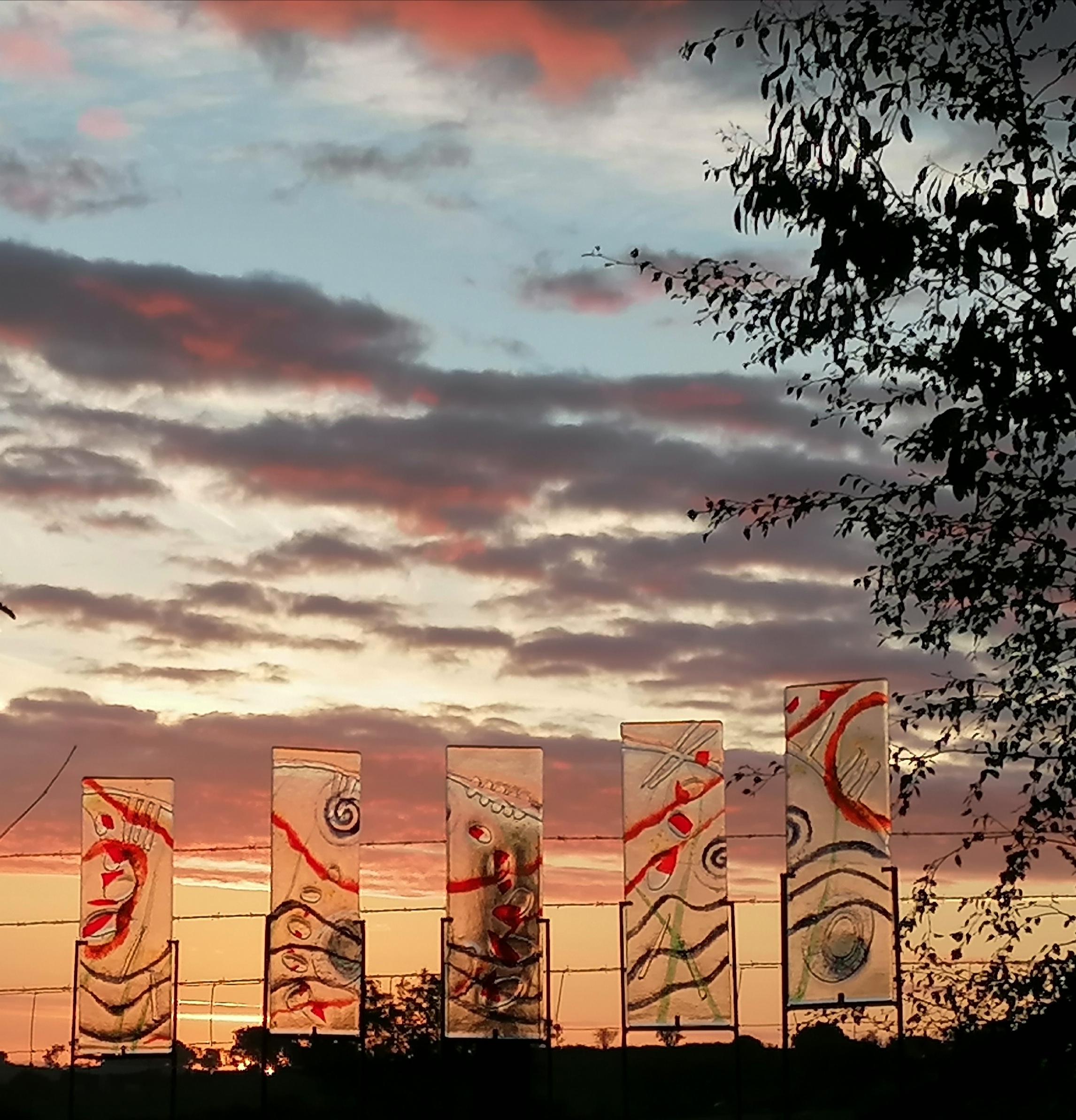 Five see through glass rectangular sculpture with red, blue and clear spirals, wavy lines and patterns on them, photographed against a pink and blue sunset sky.